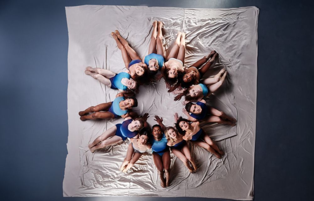 A group of ballerinas sit on the ground on top of a sheet forming a circle.