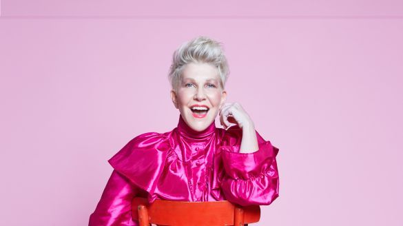 Joyce DiDonato smiling with her mouth open as she rests her hand on top of a blood orange chair.
