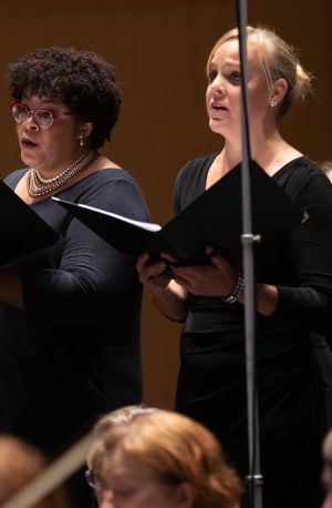 A White woman with straight dark hair, a Black woman with curly dark hair, and a White woman with blond hair in a bun sing in a chorus