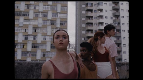 Four dancers stand close together in a diagonal row on a rooftop, tall buildings flanked behind them in a gray sky. Female dancers wear solid red or gold tops, and one male dancer wears a cream button down. One dancer looks at the camera while the others look  to the sides. 