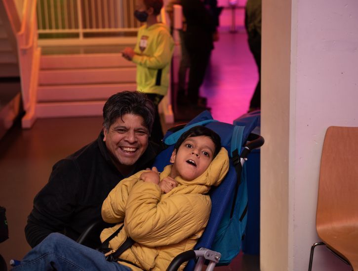 Father wears a black coat and son wears a yellow jacket, sitting in a blue wheelchair, smiling for the camera.
